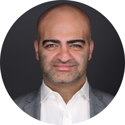Head-and-shoulders portrait of a bald man with light stubble, wearing a white shirt and gray blazer, smiling softly.
